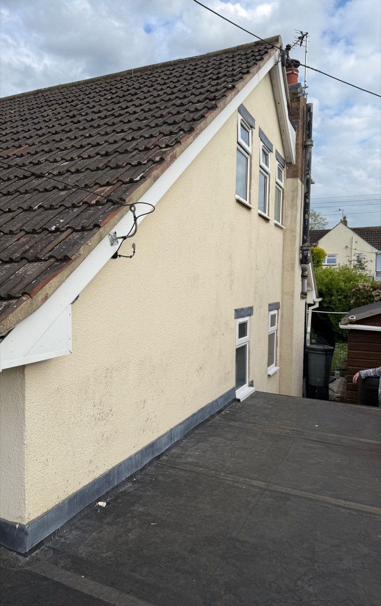The gable end of a bungalow in tired cream render, stained and patched beneath a brown pantile roof